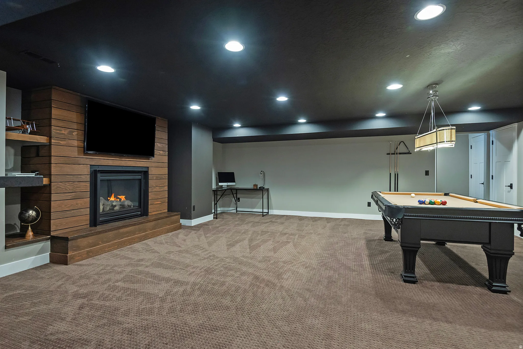 Expansive recreation room featuring a wood-finish accent wall with integrated gas fireplace and flat-screen television mount, complemented by recessed lighting throughout