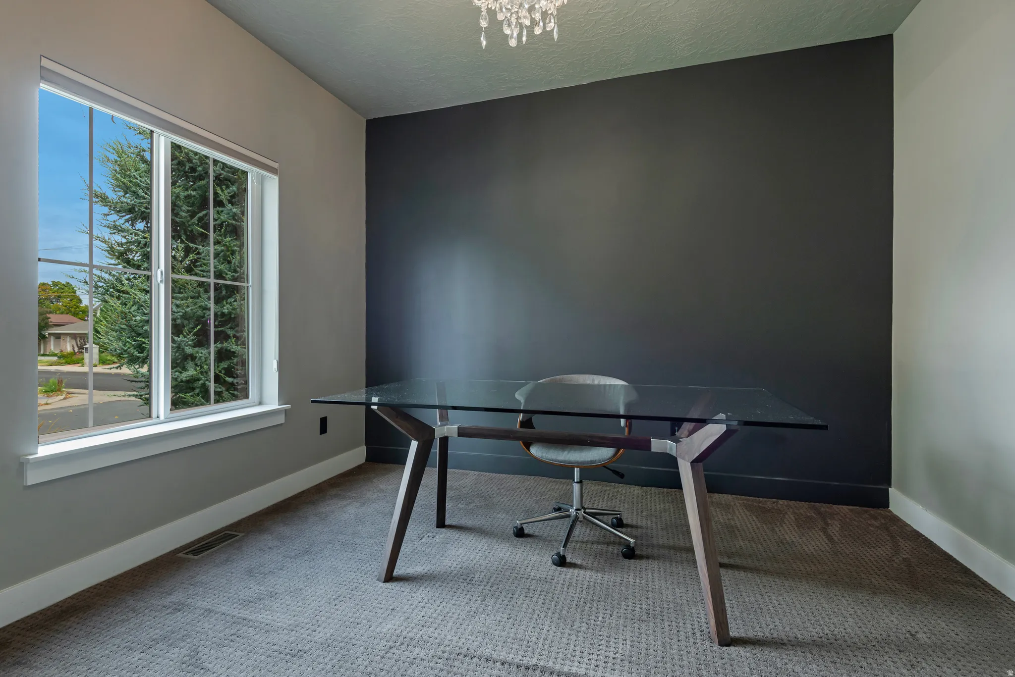 Modern home office featuring a sleek glass-top desk, neutral wall palette with a dramatic charcoal accent wall, light-filled large windows with natural outdoor sightlines, plush area rug, and contemporary crystal chandelier