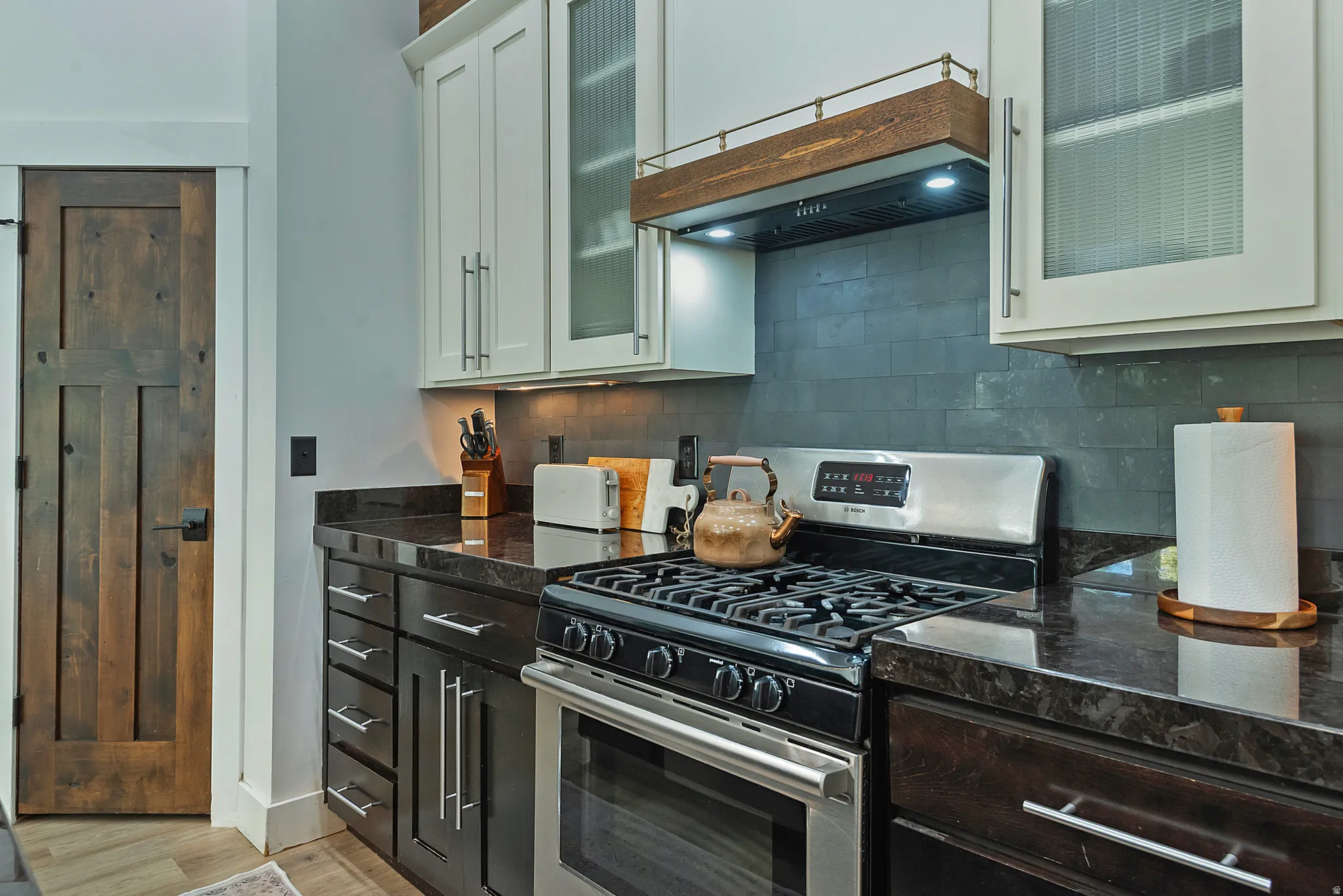 Chef's kitchen featuring professional-grade gas range with multiple burners and convection oven, black quartz countertops, white cabinetry with glass-front upper cabinets, stainless steel appliances, tile backsplash, and under-cabinet lighting
