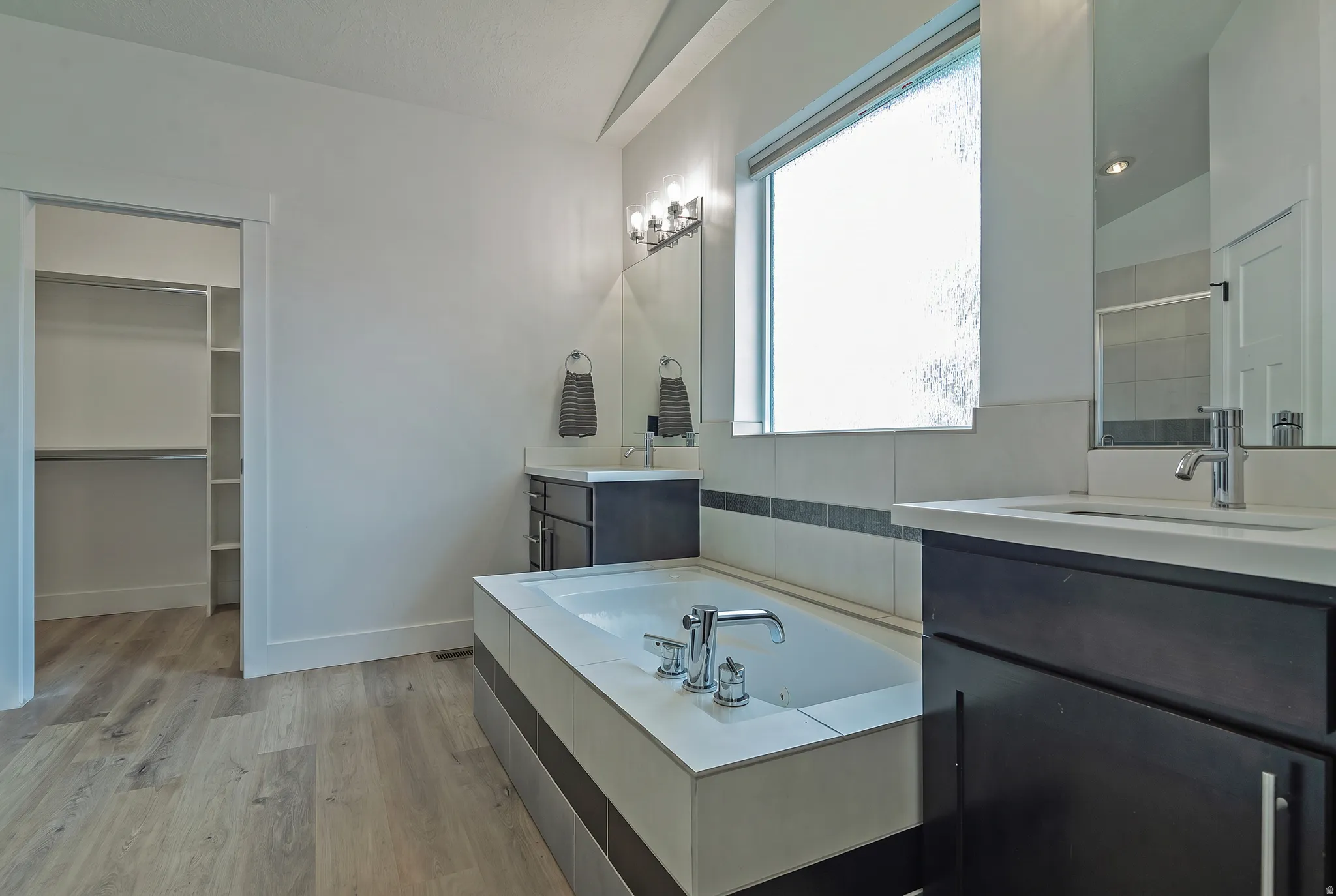 Spa-inspired primary bathroom featuring a soaking tub with chrome fixtures, dual vanity with dark cabinetry and quartz countertop, large framed mirror with integrated lighting, and wood-finish flooring