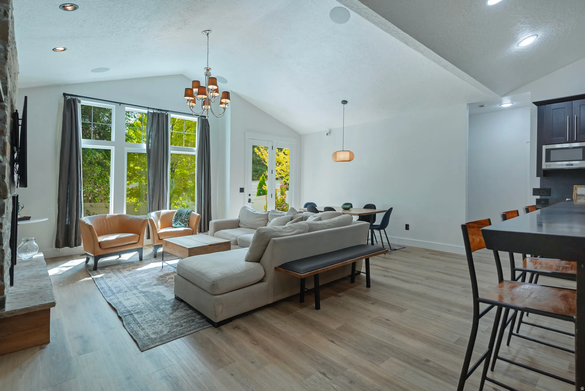 Open-concept living space featuring soaring vaulted ceilings with recessed lighting, expansive multi-pane windows with gray drapery, wide-plank wood-finish flooring, and a modern kitchen with dark cabinetry and stainless steel appliances