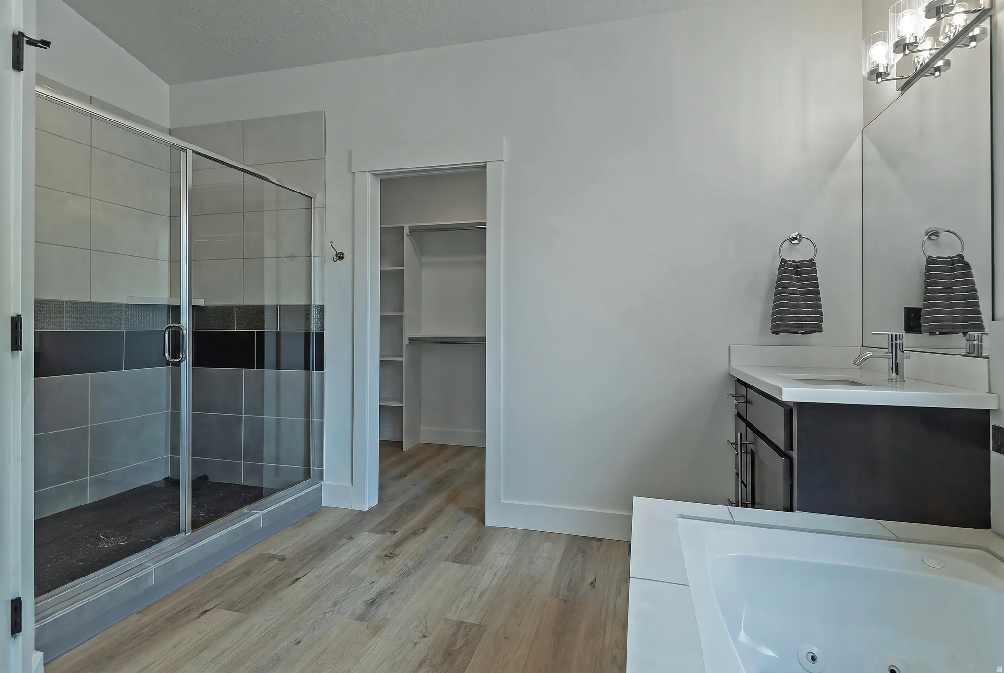 Glass-enclosed shower with tile surround, wood-finish flooring, corner soaking tub, vanity with sink, and integrated closet shelving