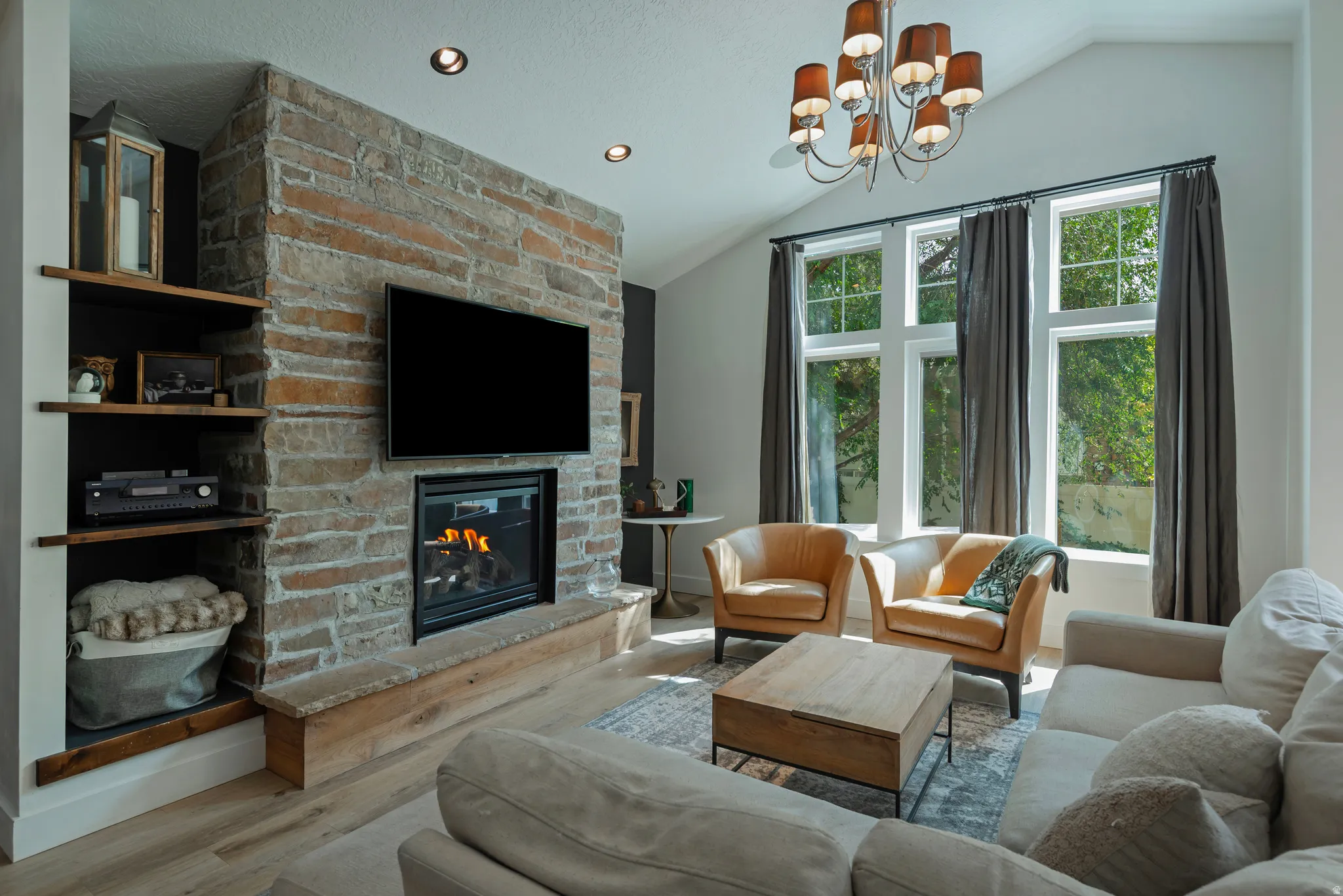 Sophisticated living room featuring a wood-finish floor, exposed brick fireplace with integrated gas insert, built-in shelving, and a statement chandelier with amber glass shades