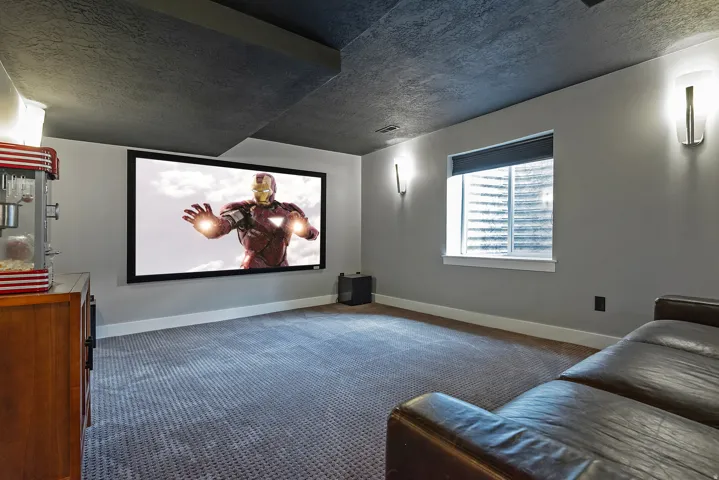 Dedicated media room featuring a fixed projection screen with surround sound capability, recessed lighting, plush seating area, polished concrete ceiling, and expansive windows with automated shades
