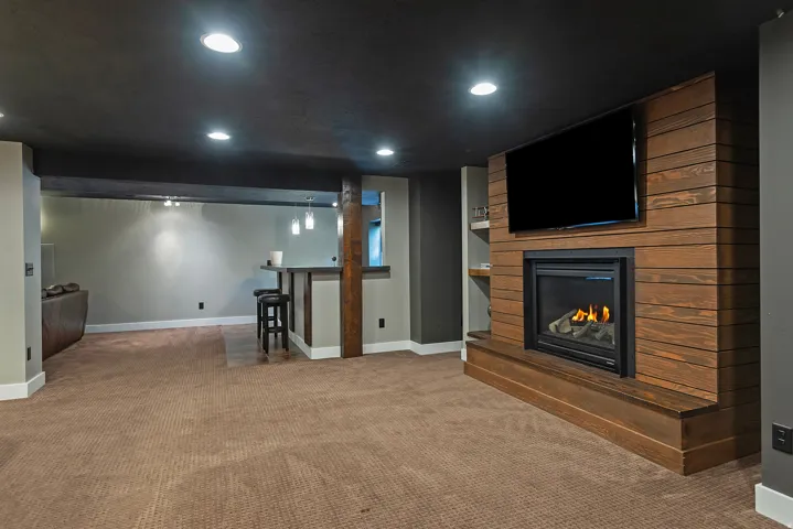 Spacious basement recreation area featuring recessed LED ceiling lighting, warm wood-finish wall paneling with an integrated gas fireplace and media nook, neutral carpet flooring, and multiple finished zones ideal for entertaining or personal use