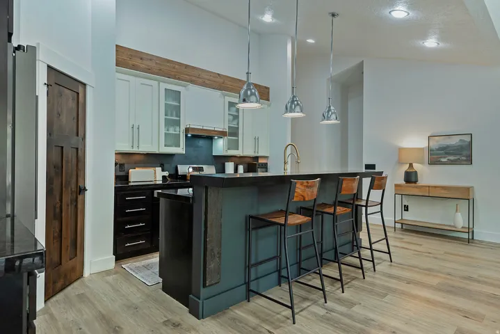 Kitchen featuring a central island with pendant lighting, white upper cabinetry with glass inserts, black lower cabinetry, wood-finish flooring, and a dark wood-paneled door