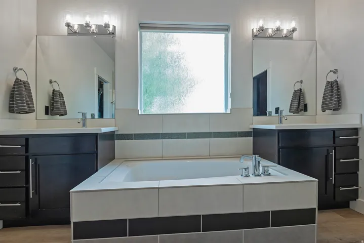 Spa-inspired primary bathroom featuring dual vanities with black cabinetry and quartz countertops, large soaking tub with tile surround, dual framed mirrors with integrated lighting, and a centered window flooding the space with natural light