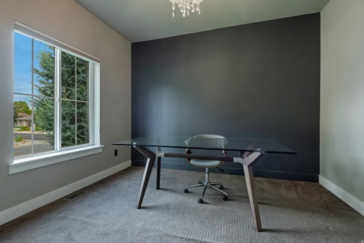 Modern home office featuring a sleek glass-top desk, neutral wall palette with a dramatic charcoal accent wall, light-filled large windows with natural outdoor sightlines, plush area rug, and contemporary crystal chandelier
