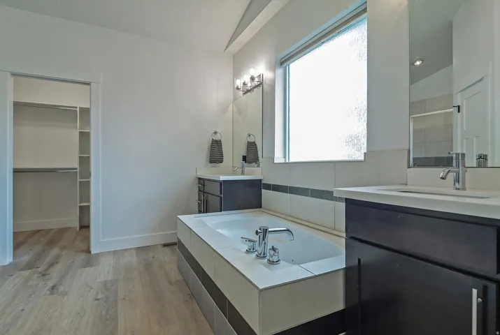 Spa-inspired primary bathroom featuring a soaking tub with chrome fixtures, dual vanity with dark cabinetry and quartz countertop, large framed mirror with integrated lighting, and wood-finish flooring