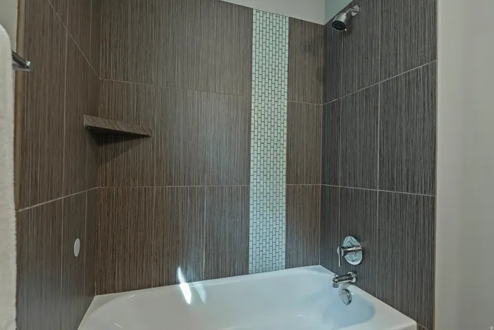 Built-in tub with chrome fixtures, wood-finish wall tiling, accent mosaic tiling, and integrated shower head
