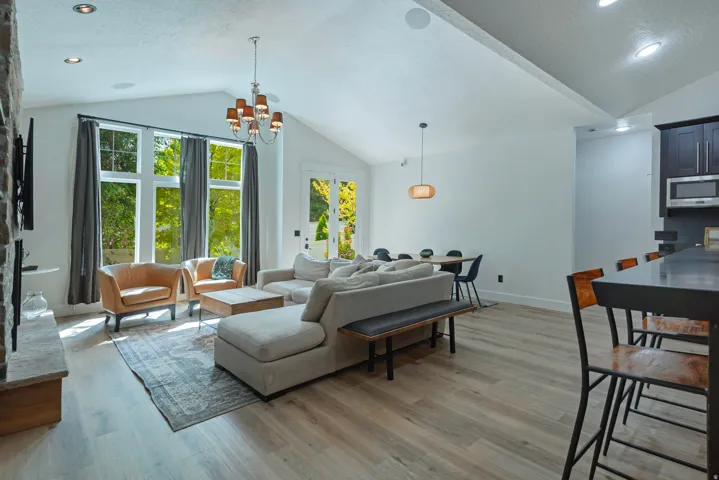 Open-concept living space featuring soaring vaulted ceilings with recessed lighting, expansive multi-pane windows with gray drapery, wide-plank wood-finish flooring, and a modern kitchen with dark cabinetry and stainless steel appliances