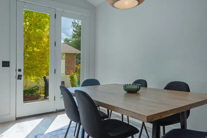 Modern dining space featuring a wood-finish table with metal base, black upholstered chairs, and abundant natural light from French doors opening to landscaped grounds