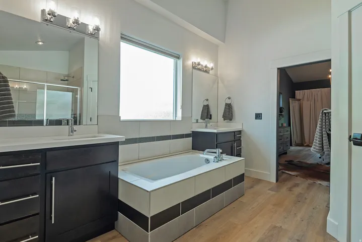 Spa-inspired master bathroom featuring dual vanities with black cabinetry and white quartz countertops, frameless mirror with integrated lighting, soaking tub with tile surround, and warm wood-finish flooring throughout