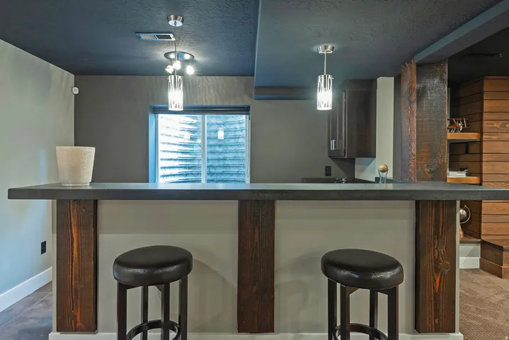 Modern wet bar featuring a dark wood-finished counter with waterfall edge, dual black leather upholstered seating, pendant lighting with glass shades, and a mirrored backsplash