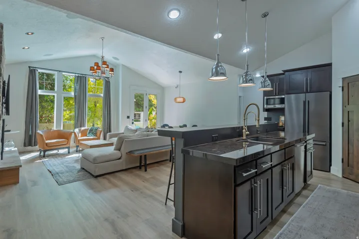 Open-concept kitchen and living area featuring a large center island with dark cabinetry, stainless steel appliances, pendant lighting, wood-finish flooring, and abundant natural light from multi-pane French doors overlooking the landscape