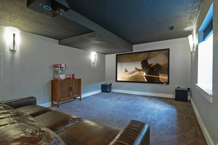 Dedicated media room featuring a recessed wall-mounted display, dark ceiling with integrated lighting, polished concrete accent beam, dual built-in speaker systems, blue geometric area flooring, and wood-finish cabinetry with storage