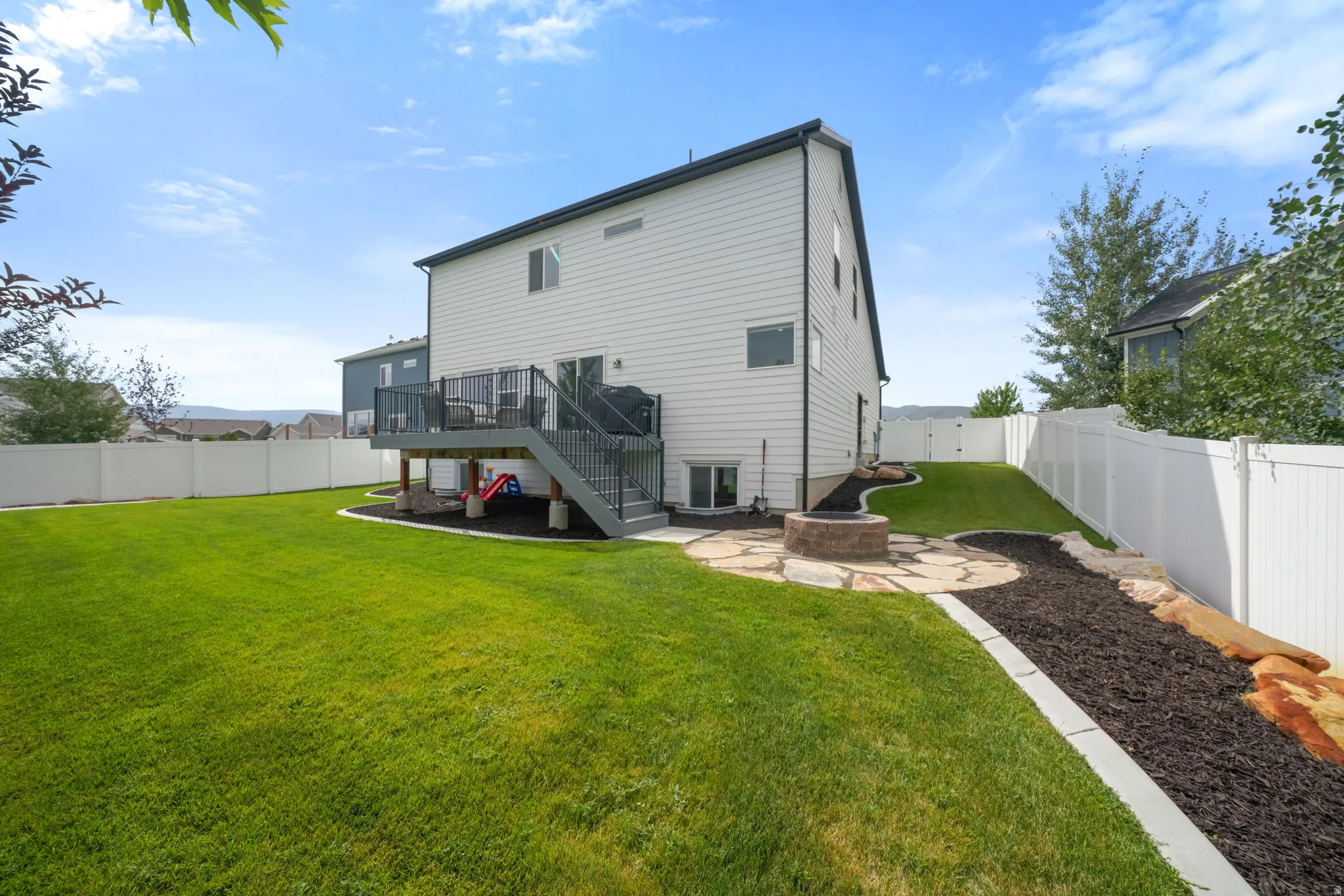 Expansive fenced backyard featuring a large green lawn, multi-level deck with stairs, a built-in fire pit with a flagstone patio, and landscaped beds with dark mulch