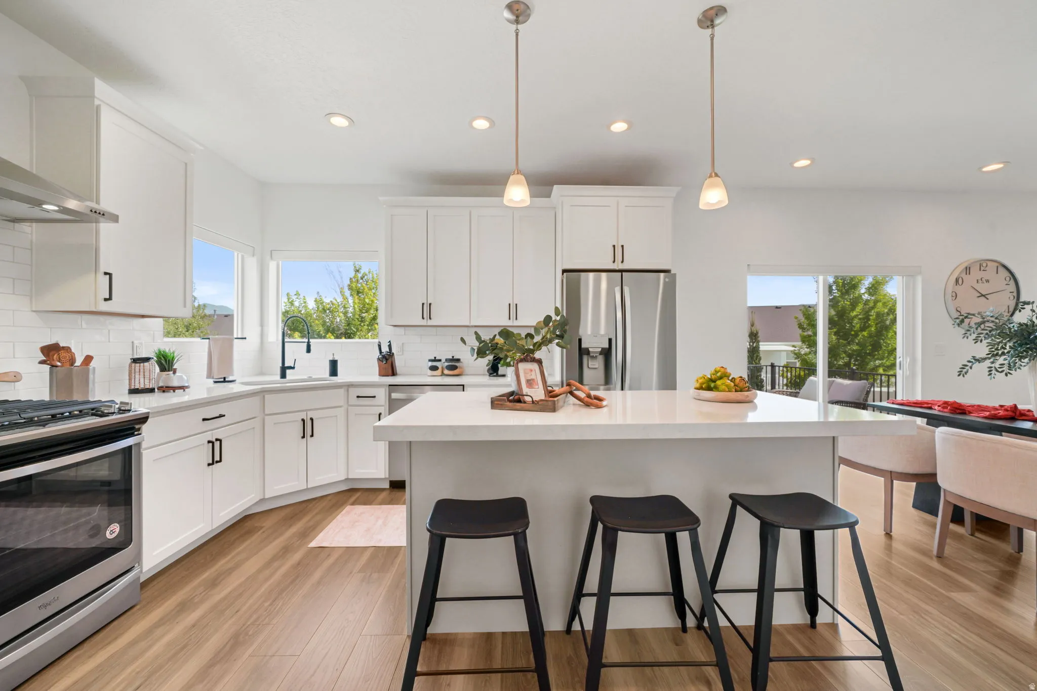 Modern kitchen featuring white cabinetry with brushed nickel hardware, a large waterfall-edge island with seating for three, stainless steel appliances including a gas range, pendant lighting over the island, and wide-plank wood-finish flooring throughout