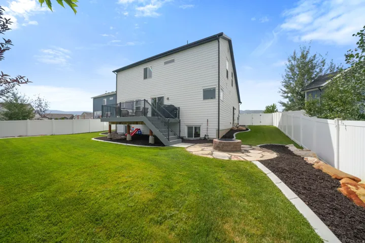 Expansive fenced backyard featuring a large green lawn, multi-level deck with stairs, a built-in fire pit with a flagstone patio, and landscaped beds with dark mulch