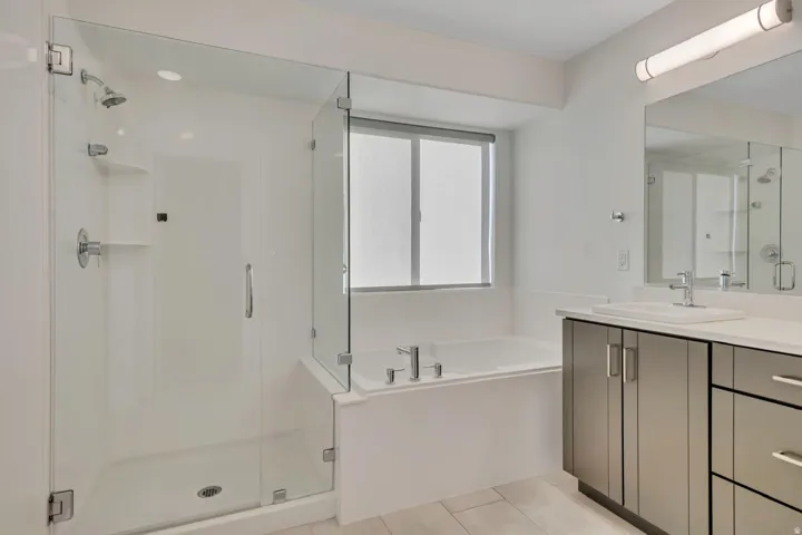 Glass-enclosed shower, soaking tub, window with roller shade, vanity with dual sinks, and light wood-finish flooring