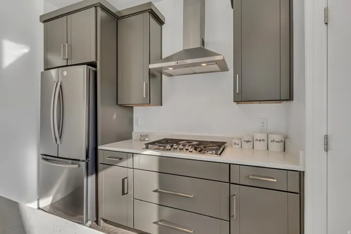 Stainless steel refrigerator, stainless steel range hood, built-in cooktop, gray cabinetry, and light-toned countertops