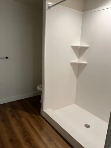 Shower enclosure with white surround, integrated corner shelving, chrome drain, and wood-finish flooring