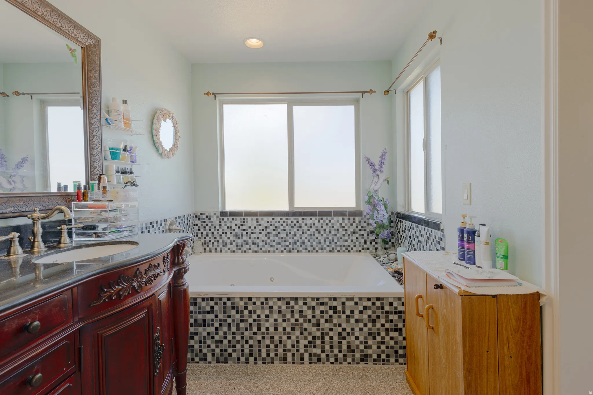 Jetted tub with mosaic tile surround, frosted windows, and a decorative vanity with an ornate mirror
