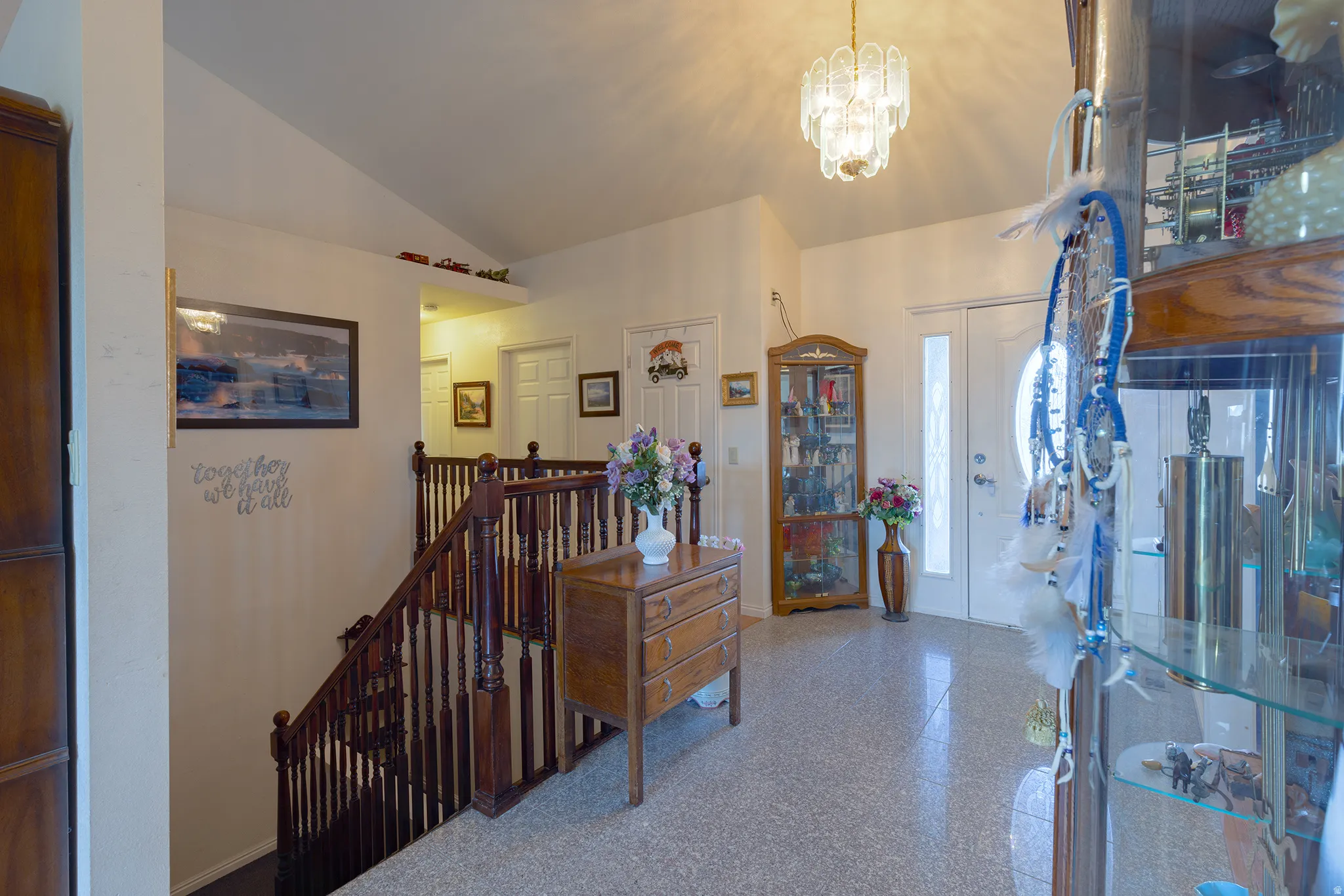 Vaulted ceiling, decorative chandelier, wood-finish staircase with balusters, light-colored flooring, and an entry door with a decorative glass insert