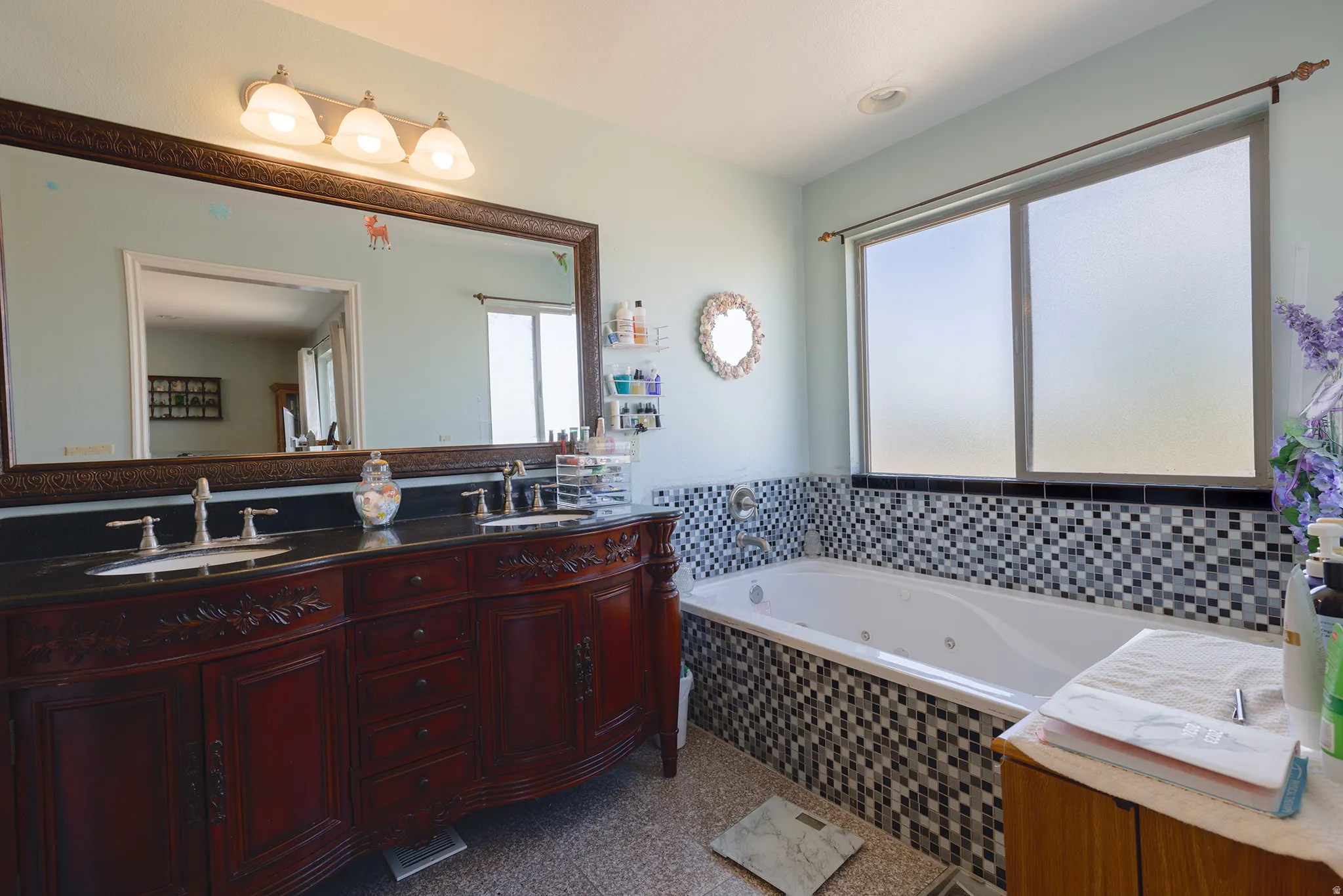 Dual-sink vanity with dark countertop, large framed mirror, built-in soaking tub with mosaic tile surround, and frosted window