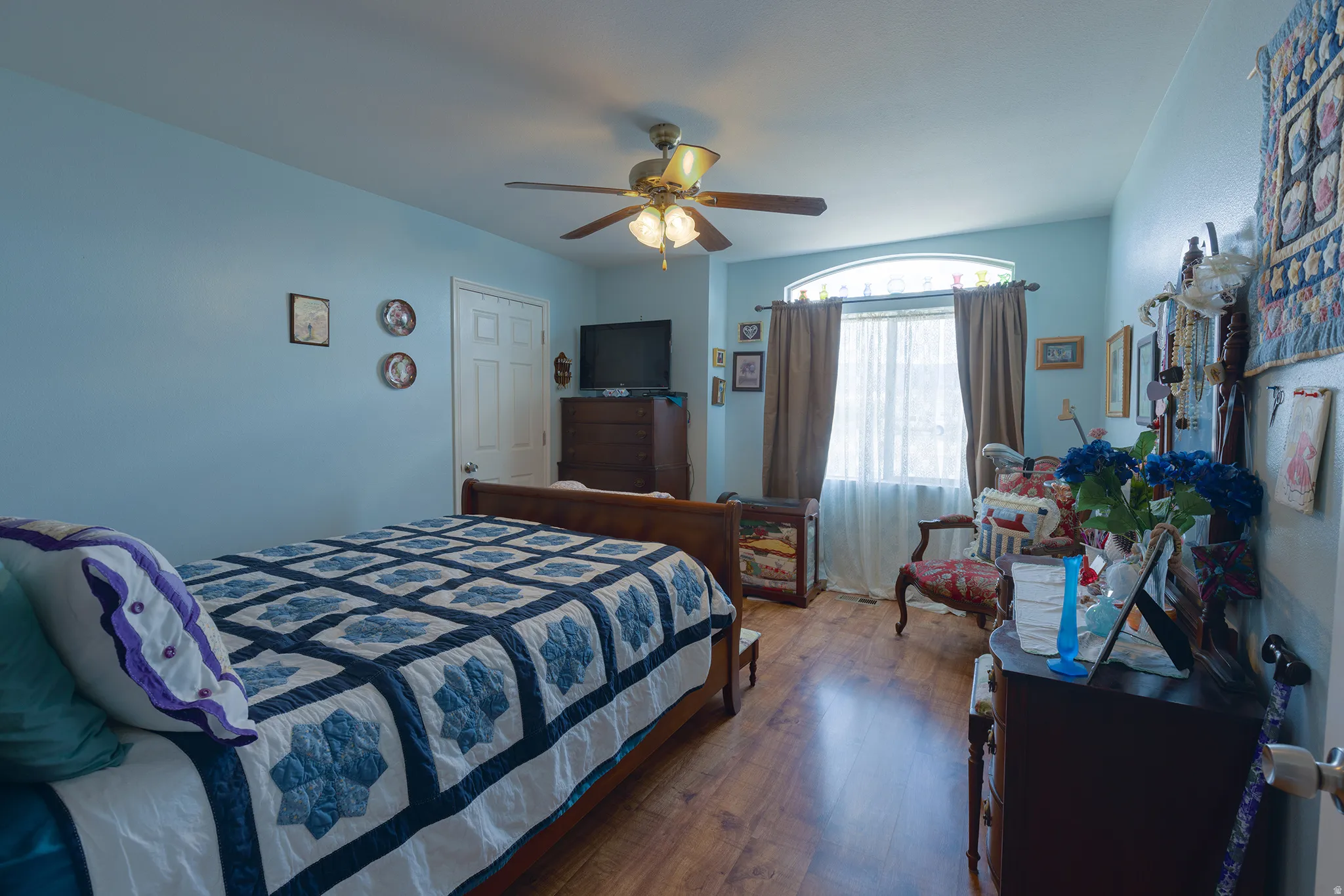 Wood-finish flooring, ceiling fan with integrated lighting, door with panel detailing, arched transom window, and window with light-filtering sheers