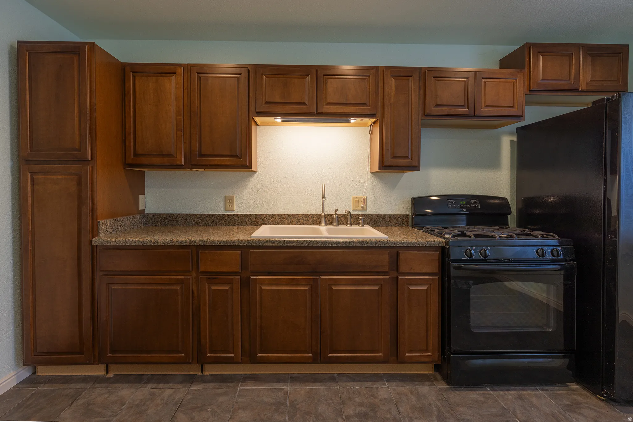 Kitchen featuring wood-finish cabinetry, a double basin sink, under-cabinet lighting, a black gas range, and a black refrigerator