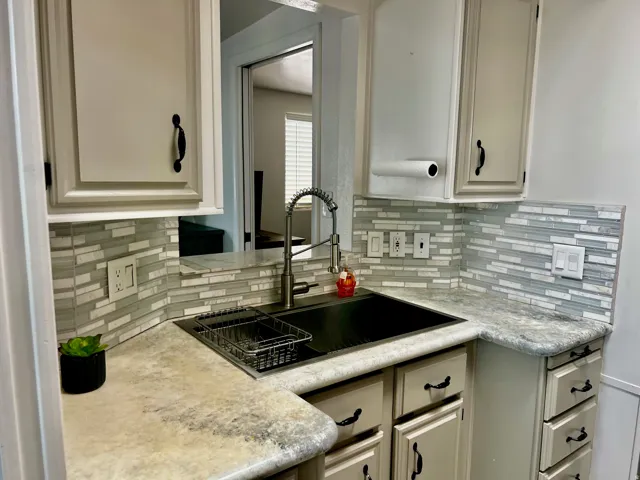 Updated kitchen featuring light-toned cabinetry with dark hardware, a tile backsplash, a dark basin sink with a flexible faucet, and a pass-through window