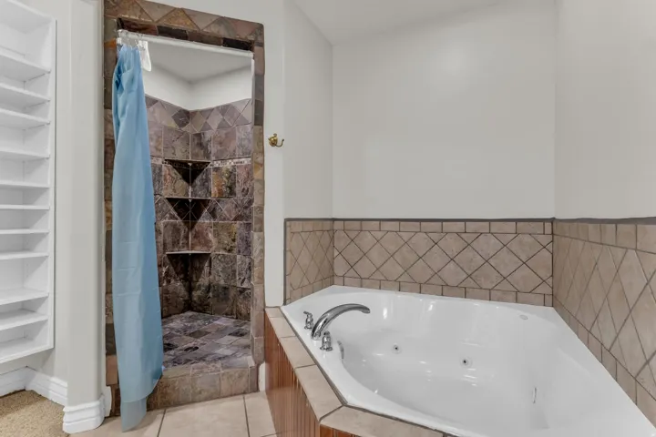 Corner soaking tub with deck-mounted fixtures, stone-tiled shower, and built-in shelving