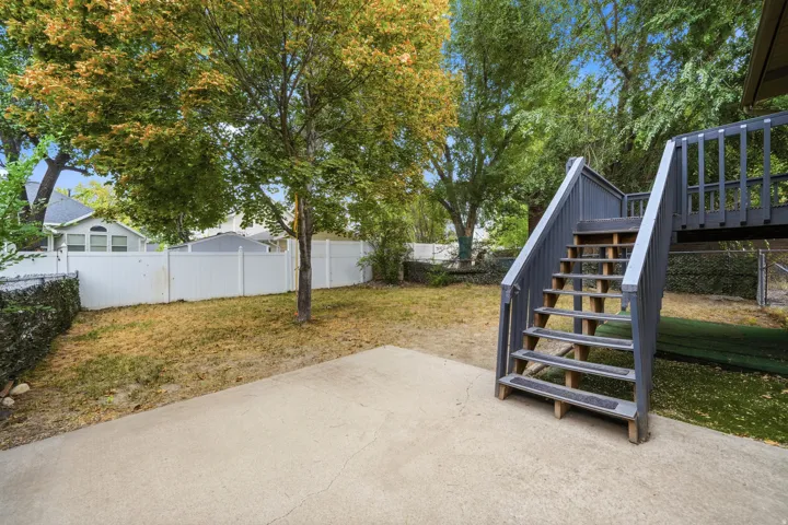 Expansive backyard featuring a concrete patio, mature trees, white privacy fencing, and a multi-level deck with stairs