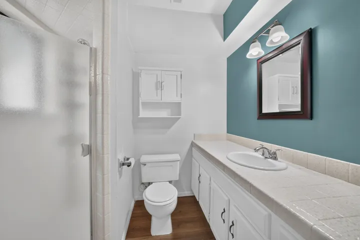 Shower enclosure with white tile trim, wood-finish flooring, white vanity with integrated sink, framed mirror, and wall-mounted light fixture