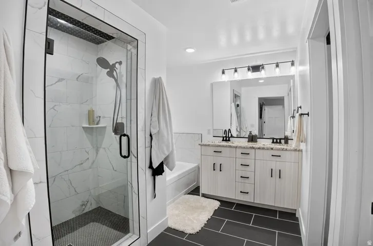 Walk-in shower with clear glass enclosure and contrasting tile, dual vanity with granite countertop and undermount sinks, separate bathtub with tiled surround, and dark large-format tile flooring