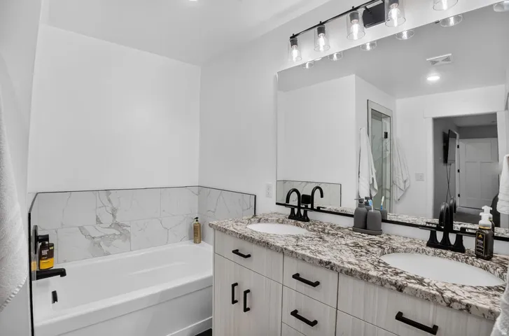 Dual vanity with undermount sinks and stone countertops, black hardware, wood-finish cabinetry, built-in bathtub with tile surround, and recessed lighting