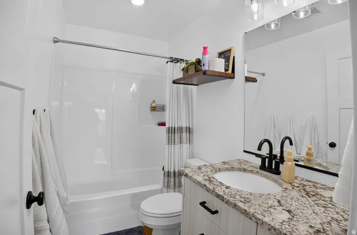 White integrated shower and tub with a built-in shelf, vanity with a stone-finish countertop, under-mount sink, and matte black faucet, and a large wall-mounted mirror