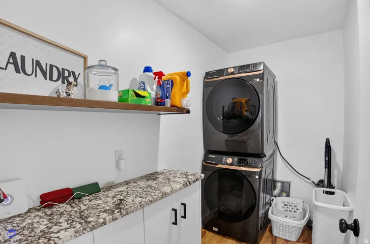 Stacked washer and dryer units, wood-finish flooring, floating wood-tone shelf, white cabinetry with dark pulls, and a granite-patterned countertop