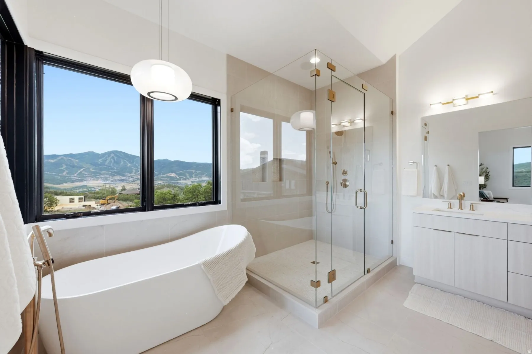 Freestanding soaking tub, glass-enclosed shower, dual vanity with undermount sink, mountain backdrop, and contemporary light fixtures