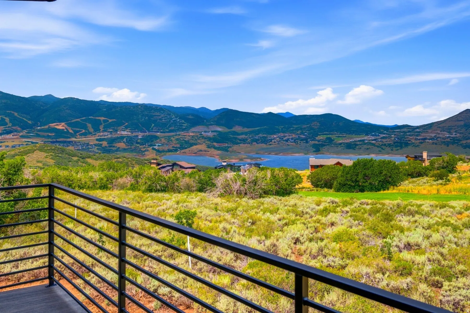 Exterior balcony with a black horizontal-bar railing, showcasing expansive mountain and lake scenery, and varied natural landscaping