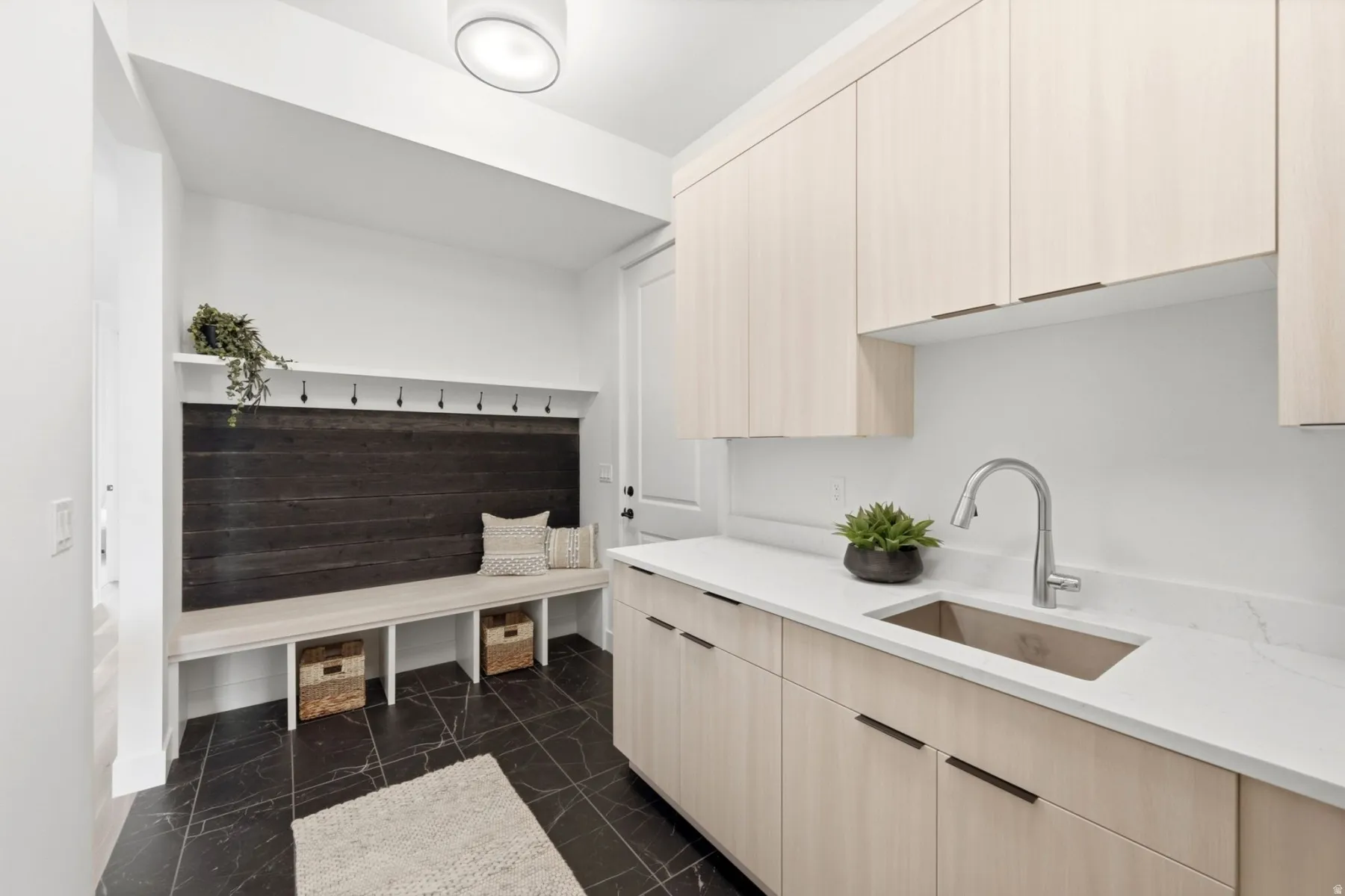 Built-in bench seating with storage, dark wood-panel accent wall, coat hooks, light wood-finish cabinetry, white countertop, and single-basin sink with a gooseneck faucet