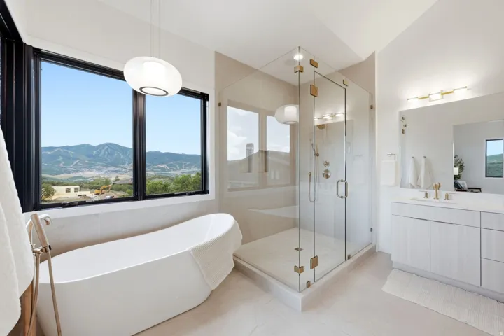 Freestanding soaking tub, glass-enclosed shower, dual vanity with undermount sink, mountain backdrop, and contemporary light fixtures