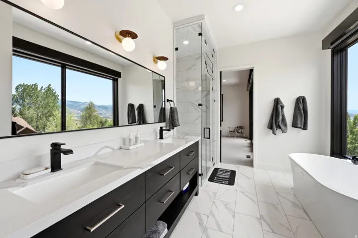 Dual vanity with integrated sinks, frameless glass shower enclosure, freestanding soaking tub, large format floor tile, and oversized windows