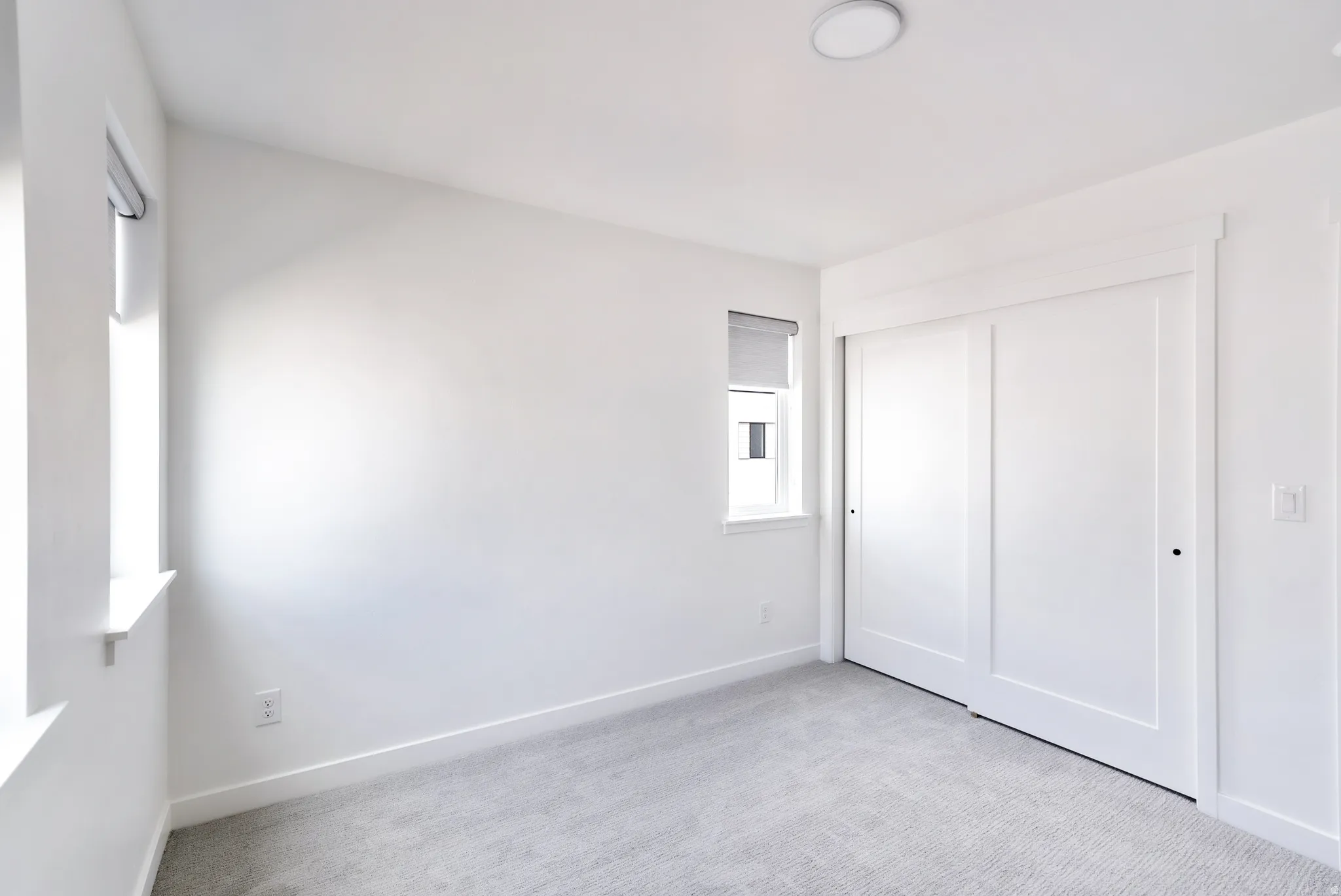 Recessed lighting fixture, two windows with roller shades, white walls, light gray carpet, and two-panel sliding closet doors