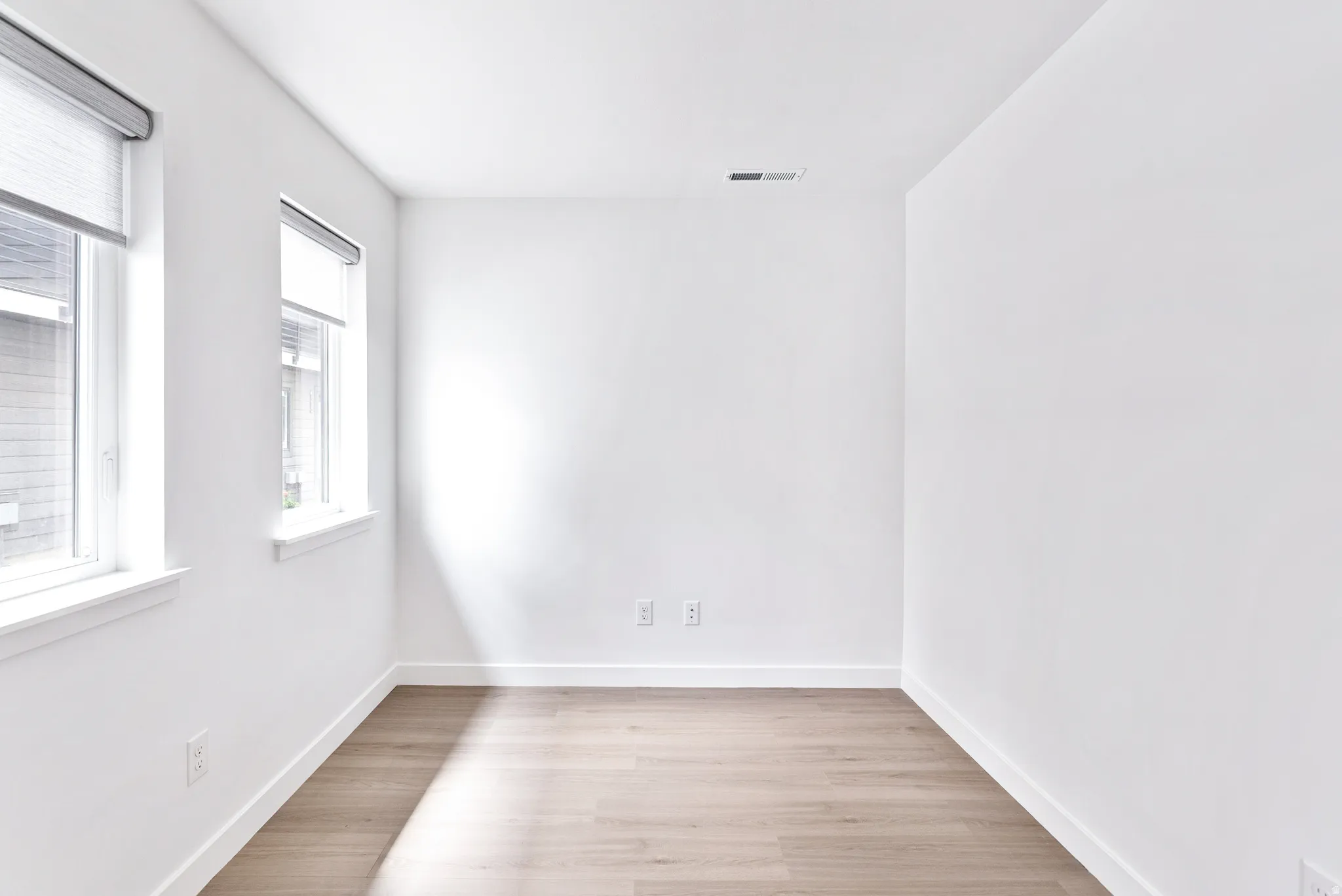 Light wood-finish flooring, white walls, and two windows with roller shades