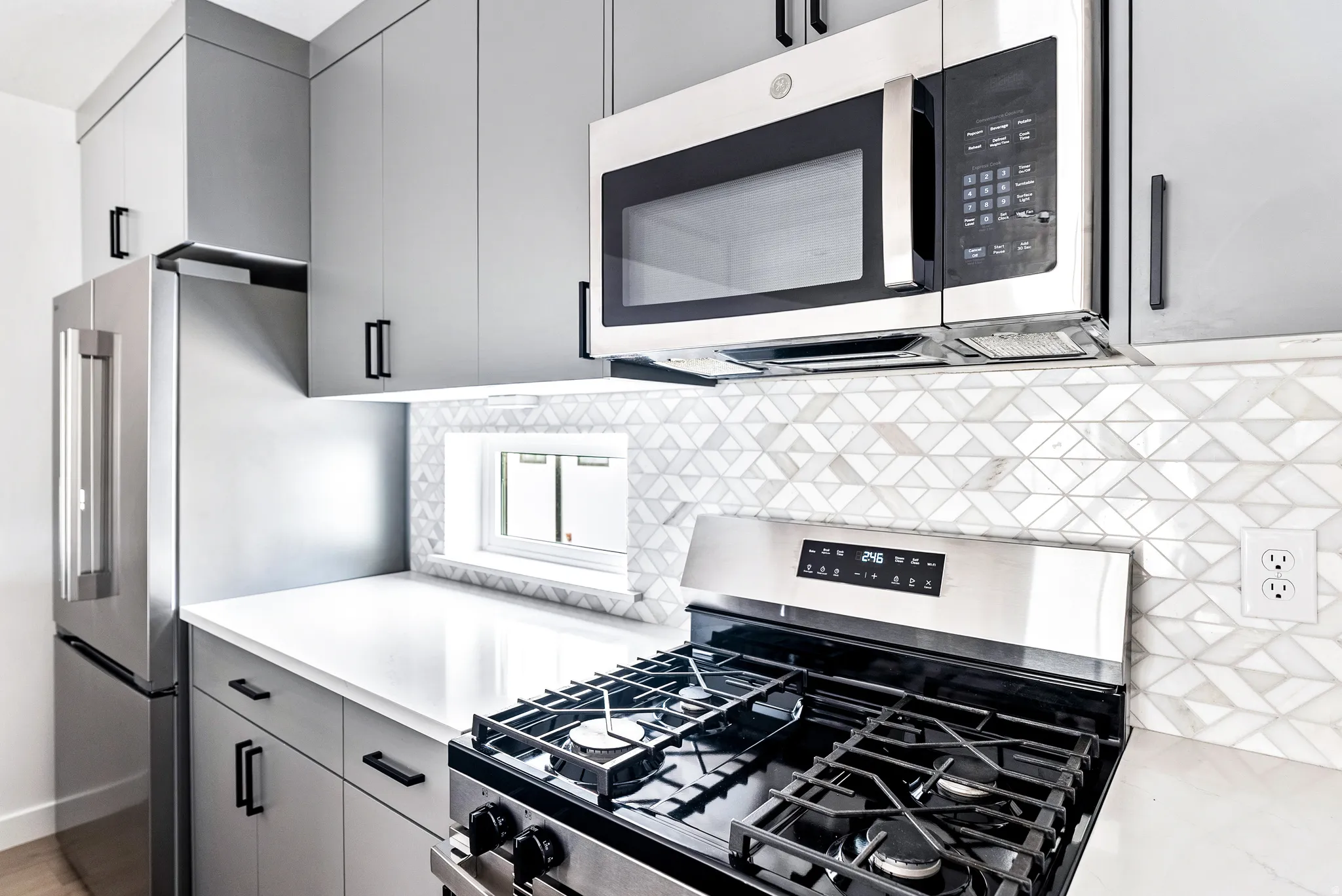 Sleek kitchen featuring gray cabinetry with black hardware, stainless steel appliances, white countertops, and a decorative tile backsplash