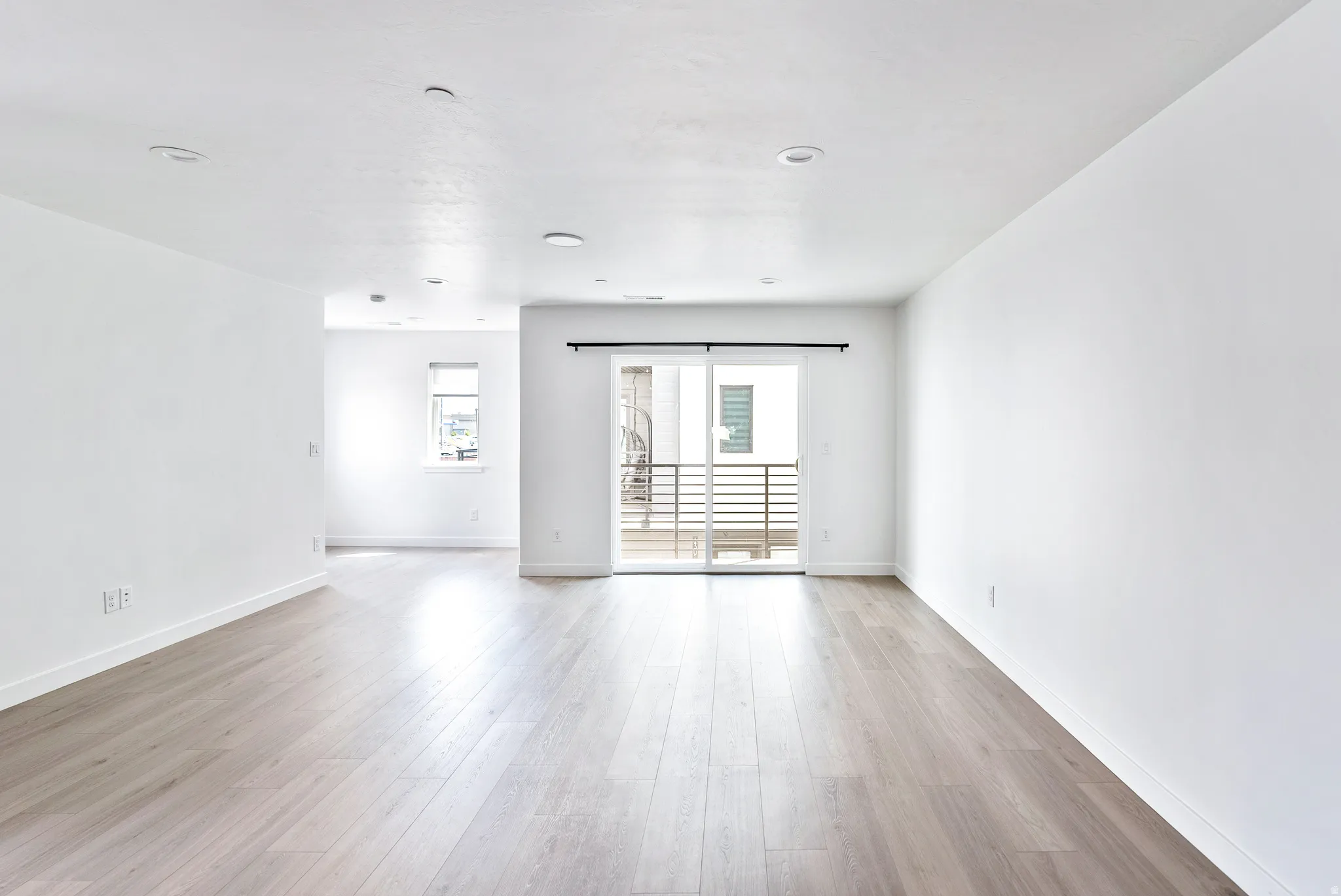 Expansive living area featuring wood-finish flooring, recessed lighting, a large window, and a sliding glass door leading to a balcony with metal railings
