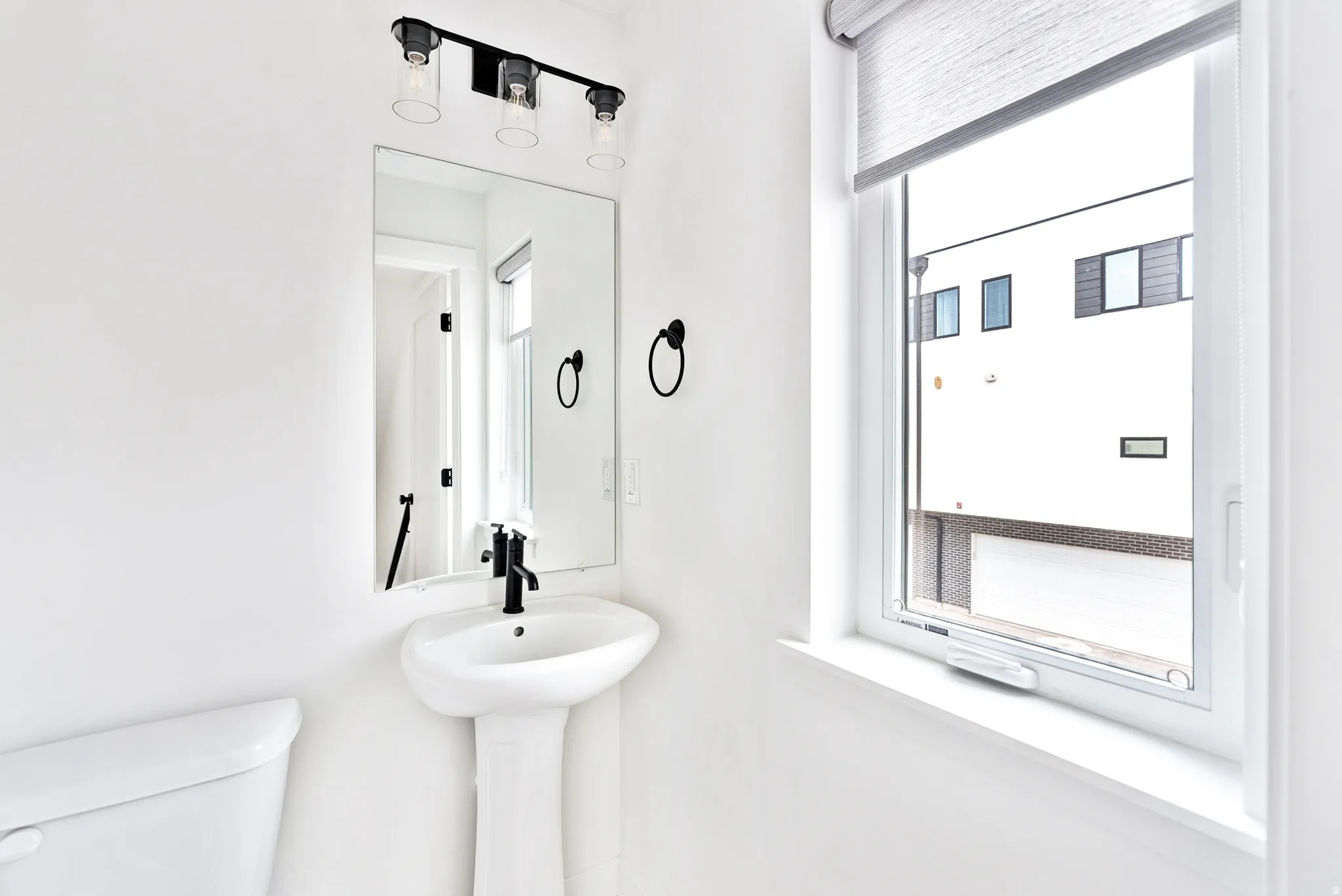 Pedestal sink with matte black faucet, wall-mounted mirror, and contemporary vanity light fixture