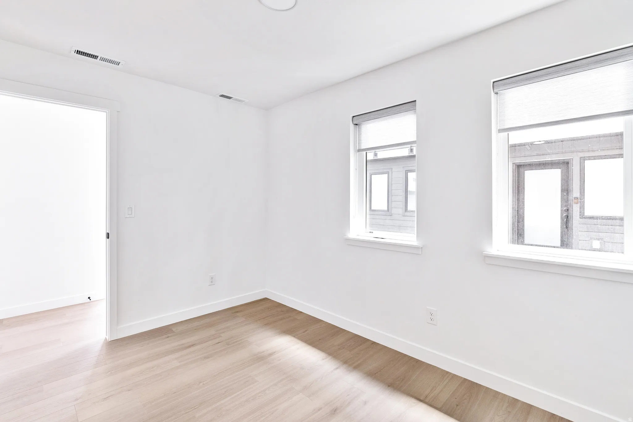 Light wood-finish flooring, two large windows, roller shades, white walls, and a recessed ceiling light
